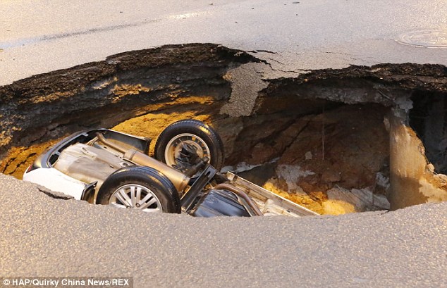 The car flipped upside down after falling into the sinkhole; the car's rear wheels can be seen just below the road surface which has broken up near a drainage hole